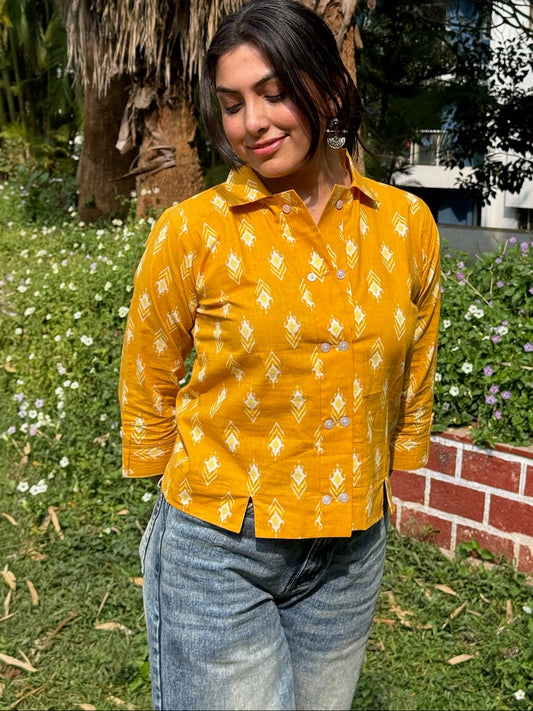 Woman wearing a yellow patterned shirt and blue jeans standing outdoors with greenery in the background